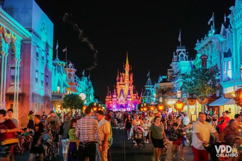 Imagem do Magic Kingdom à noite, com muitos visitantes e funcionários. O castelo e as lojas estão iluminados por luzes coloridas. 