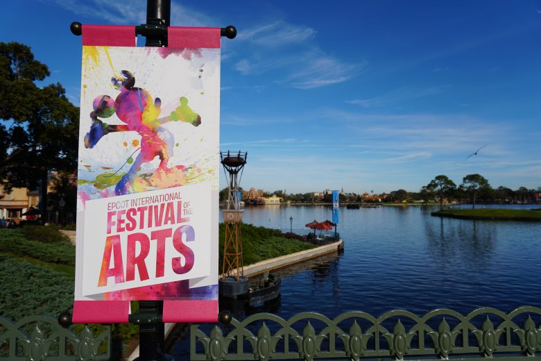 Foto da placa do Epcot International Festival of the Arts, com o Mickey pintado em estilo de aquarela, com o lago do parque ao fundo 