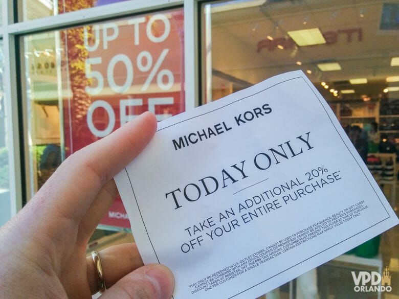 O feriado de fevereiro traz lotação e bons descontos nos outlets! Foto de uma mão segurando um cupom de desconto para a loja Michael Kors, anunciando 20% de desconto 