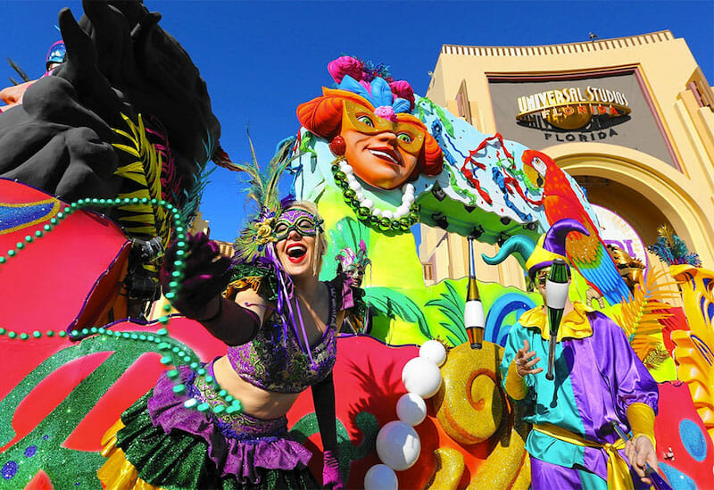 A imagem mostra várias pessoas fantasiadas com roupas e adereços bem coloridos, em frente ao arco da Universal. O Mardi Gras 2021 acontece entre fevereiro e março.