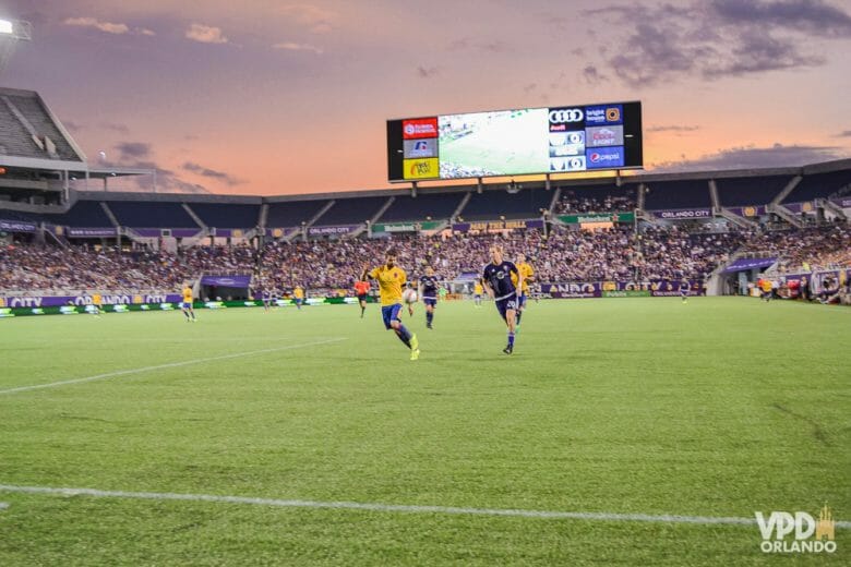 Em março começam as partidas de futebol do Orlando City! Foto do campo durante o jogo de futebol. É possível ver a arquibancada, o telão e alguns jogadores. 