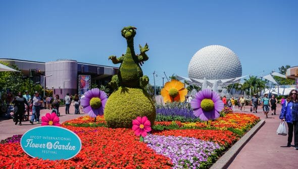 Foto do Flower & Garden no Epcot. É possível ver o céu azul, a bola do Epcot e muitas flores coloridas, assim como uma topiária com a forma do dragão da Malévola.
