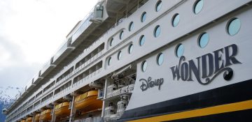 Foto do navio Disney Wonder, que faz parte da frota da Disney Cruise Line. É possível ver o nome do navio escrito em preto na lateral, as janelas redondas e detalhes em amarelo sobre a pintura.