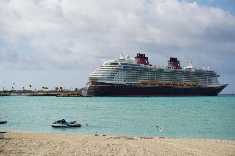 Foto do navio de cruzeiro da Disney Cruise Line perto da praia no Caribe, com o mar azul-turquesa, a areia clara e o céu claro atrás. 