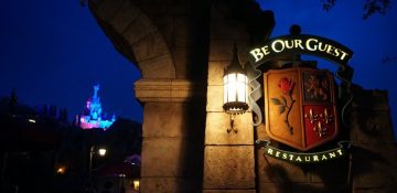 O castelo fica ainda mais bonito a noite. Foto da placa na entrada do restaurante no Magic Kingdom, durante a noite, com o castelo da Cinderela no fundo da foto. A placa iimita um brasão medieval com uma rosa, um leão e espadas.