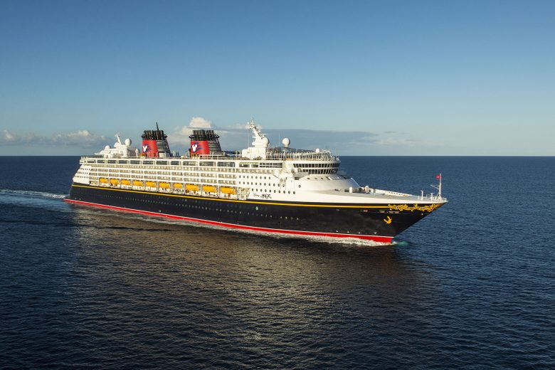 Imagem do navio de cruzeiro da Disney, o Disney Magic, em alto mar, com o céu azul ao fundo. Ele tem detalhes em amarelo e vermelho. 