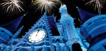 Foto do castelo da Cinderela iluminado em azul durante o show de fogos de Ano Novo no Magic Kingdom. É possível ver alguns fogos no céu acima do castelo.