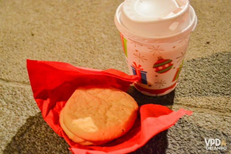 Foto de um copo branco decorado de Natal com chocolate quente e dois cookies de açúcar em um guardanapo vermelho.