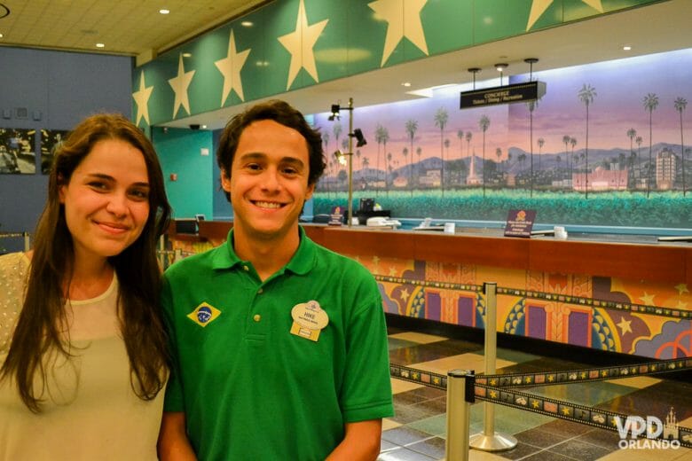 Foto da Bia e do Hike, quando eles trabalharam como Super Greeter no hotel All Star. A Bia está de roupa branca e o Hike está com o uniforme verde do hotel. 