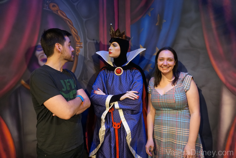 Foto da Renata e do Felipe com a Rainha Má da Branca de Neve no Storybook Dining. O Felipe está de braços cruzados encarando a Rainha e ela está encarando de volta. 