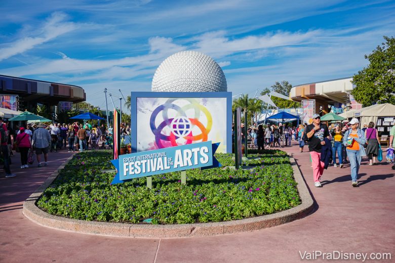 Foto de uma placa anunciando o International Festival of the Arts no Epcot, com a famosa "bola" do parque e o céu azul ao fundo