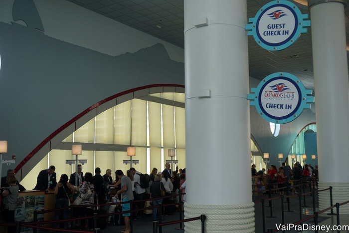 Há uma fila de check-in para quem está indo pela primeira vez (guest) e outra para quem já fez outros cruzeiros (castaway club members). Foto de visitantes na fila para fazer check-in no navio da Disney, com duas placas indicando os locais. 