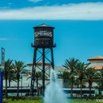 Foto da caixa d'água de Disney Springs, que leva o nome do local, com as árvores ao redor, uma fonte espirrando água mais embaixo e o céu azul ao fundo.