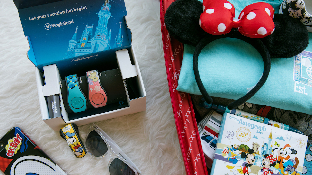 Existe também o opcional de pagar um pouco a mais por uma MagicBand mais elaborada, com desenhos e personagens. Foto de alguns itens sobre a cama: três MagicBands personalizadas, orelhinhas da Minnie, uma capinha de celular personalizada e óculos de sol. 