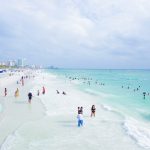 Foto da praia de Clearwater, na Flórida, mostrando a areia, o mar transparente, o céu claro e alguns visitantes passeando