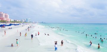 Foto da praia de Clearwater, na Flórida, mostrando a areia, o mar transparente, o céu claro e alguns visitantes passeando