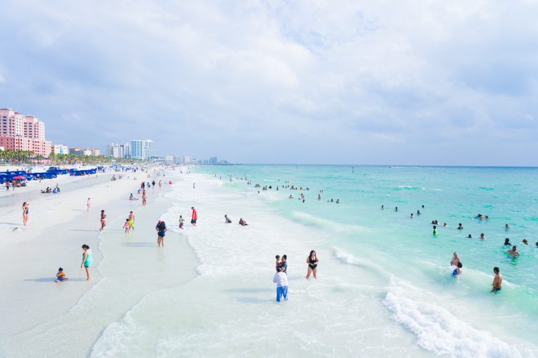 Foto da praia de Clearwater, na Flórida, mostrando a areia, o mar transparente, o céu claro e alguns visitantes passeando 