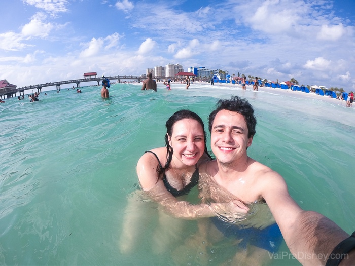 Foto da Renata e do Henrique no mar, com a areia e as pessoas ao fundo 