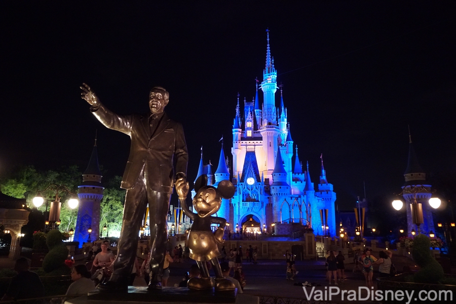 Foto do Magic Kingdom à noite, mostrando a estátua do Walt Disney com o Mickey e o castelo iluminado em azul mais ao fundo 