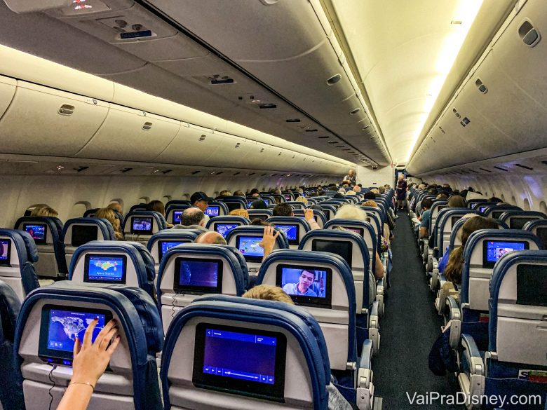 Imagem do interior de um avião. É possível ver a parte de trás de diversas fileiras de poltronas, com as telas acopladas atrás.