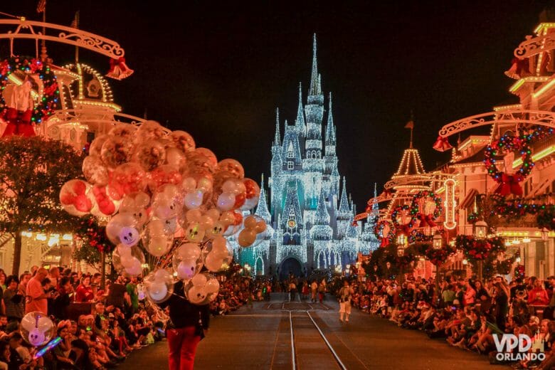 Imagem da Main Street toda decorada para o Natal. Há pessoas sentadas dos dois lados aguardando a parada e o castelo iluminado está centralizado ao fundo.