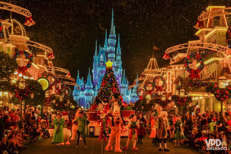 Parada de Natal da festa do Magic Kingdom. O castelo iluminado está ao fundo, a rua está decorada com guirlanda e vários personagens dançam na rua.