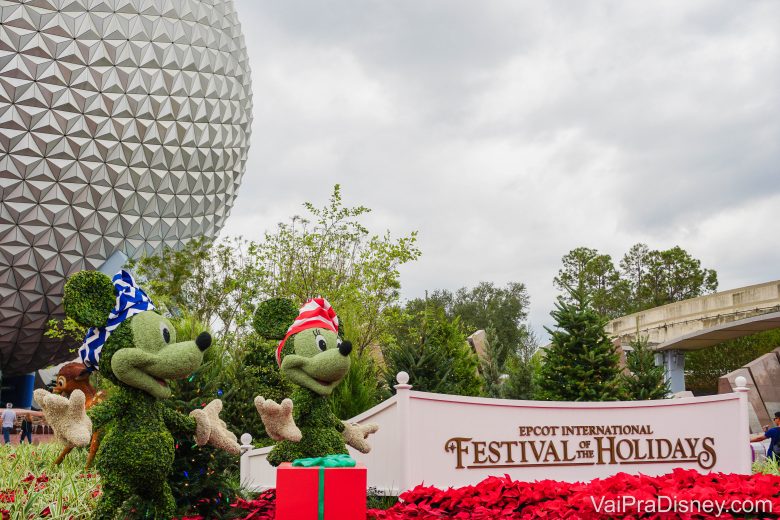 Entrada do Festival of the Holidays no Epcot. Duas topiárias do Mickey e da Minnie estão em frente à bola do Epcot,com chapéuzinhos temáticos e caixas de presente. O Festival of the Holidays 2020 acontece entre 27 de novembro e 30 de dezembro.