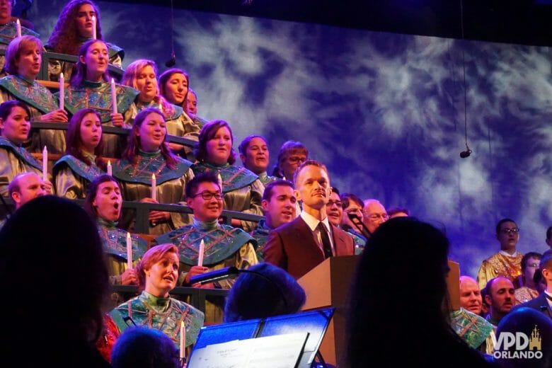 Imagem da cantata de Natal do Epcot. Várias pessoas cantam e seguram velas ao fundo, e o narrador Neil Patrick Harris está na frente.