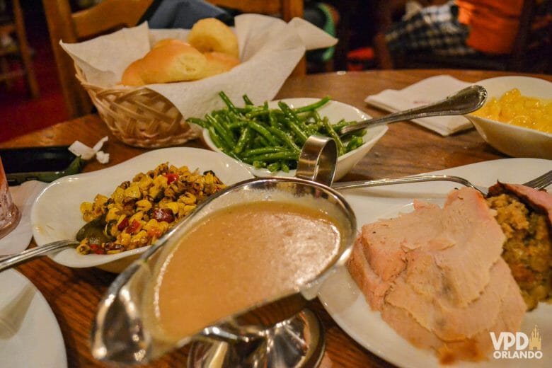 Varias comidas típicas de Natal em pratos brancos em uma mesa: há uma espécie de farofa, peru, molho, pãezinhos e vagem.