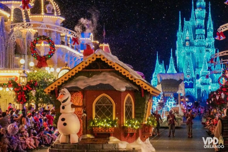 Parada de Natal do Magic Kingdom. O Olaf está em um carro que possui uma casinha coberta de Neve. O castelo iluminado está ao fundo.