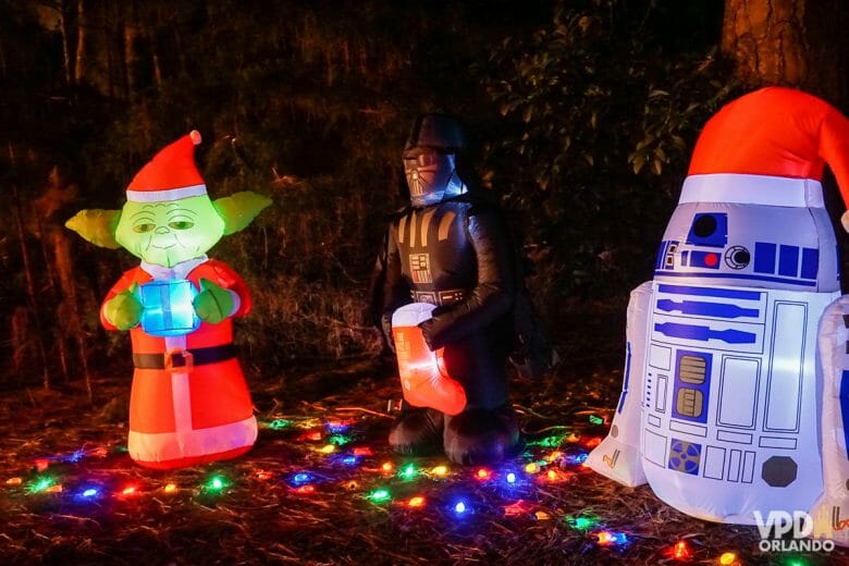 Yoda, Darth Vader e R2-D2 infláveis com chapéus e decorações de Natal. Há várias luzinhas coloridas no chão.
