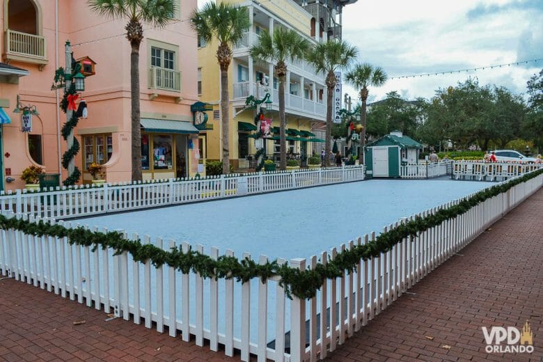 Pista de patinação no gelo sendo montada. Há várias casinhas coloridas em volta com guirlandas e decorações de Natal.