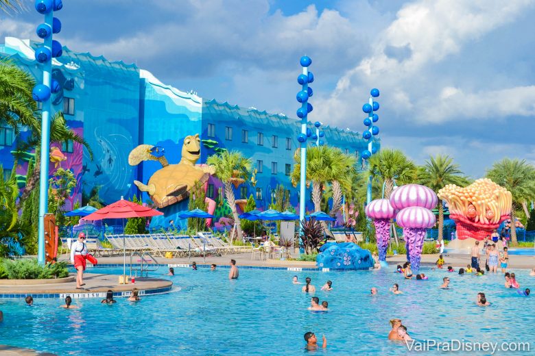 A imagem mostra visitantes na piscina do hotel da Disney, o Art of Animation. A decoração é colorida, o prédio ao fundo é azul e a tartaruga do filme Procurando Nemo é parte da decoração. 