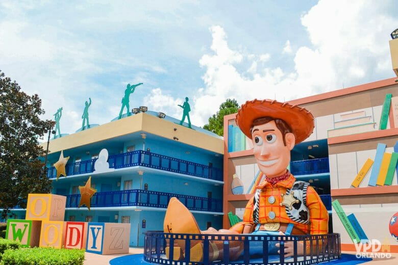 Foto do hotel Disney's All Star Movies, da área do Toy Story. Há uma réplica do Woody gigante rodeado de brinquedos, ao lado do prédio, que é azul. 