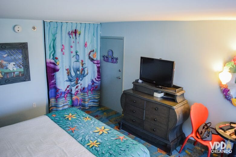 Foto do interior do quarto da Pequena Sereia no Art of Animation. É possível ver a cama e a cortina, com decorações de fundo do mar, a TV sobre um móvel, uma cadeira laranja e um quadro na parede. 