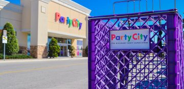 Foto do exterior da loja Party City em Orlando, com seu letreiro multicolorido ao fundo e um carrinho de compras roxo em primeiro plano, com o mesmo letreiro em versão menor