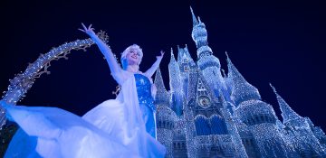 Foto da rainha Elsa em frente ao castelo da Cinderela no Magic Kingdom, iluminado em tons de azul contra o céu escuro.