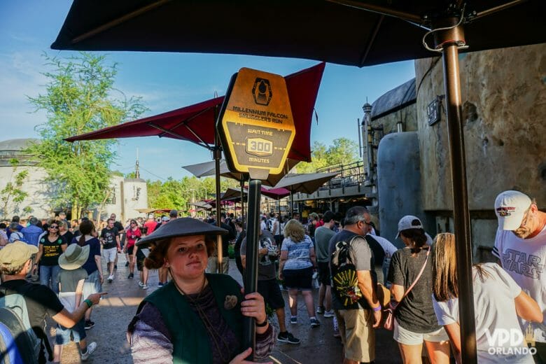 Foto de muitos visitantes em um dia de parque cheio no Hollywood Studios, formando longas filas. Uma funcionária carrega uma placa com o tempo de espera da atração. 