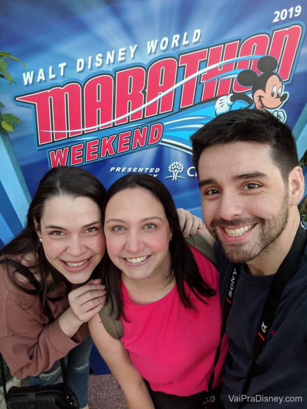 Foto da Bia, Renata e Felipe sorrindo em frente ao cartaz da maratona da Disney 