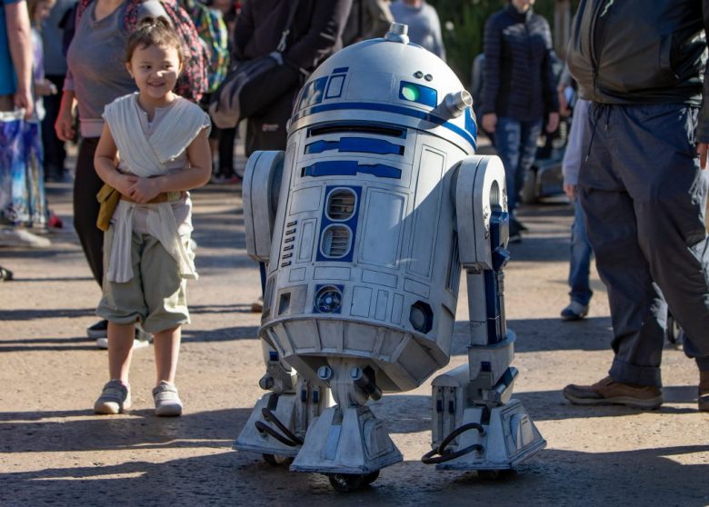 Foto do R2-D2 andando pelo parque na Califórnia, com uma menininha vestida de Rey ao lado 