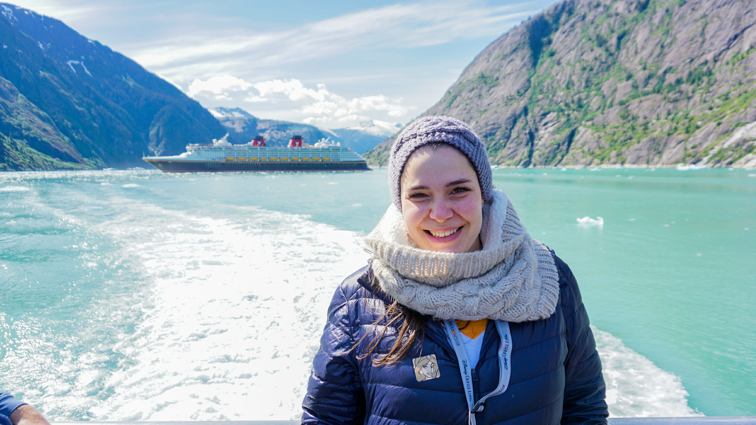 Foto da Bia com a paisagem do Alaska ao fundo. É possível ver o mar, uma montanha e o navio do cruzeiro da Disney