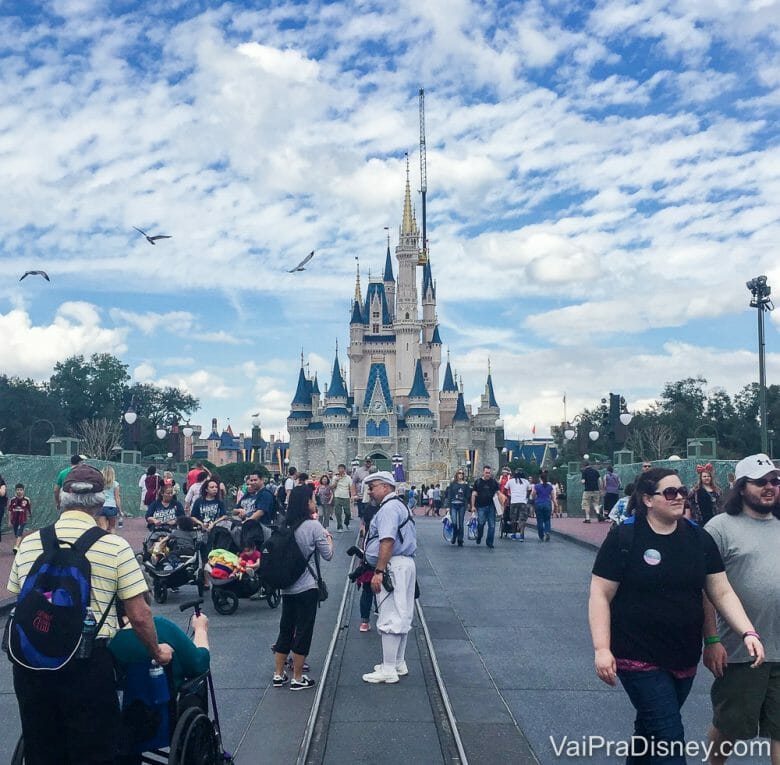 Foto do castelo da Cinderela no Magic Kingdom, com visitantes passando em frente e o céu azul ao fundo. 