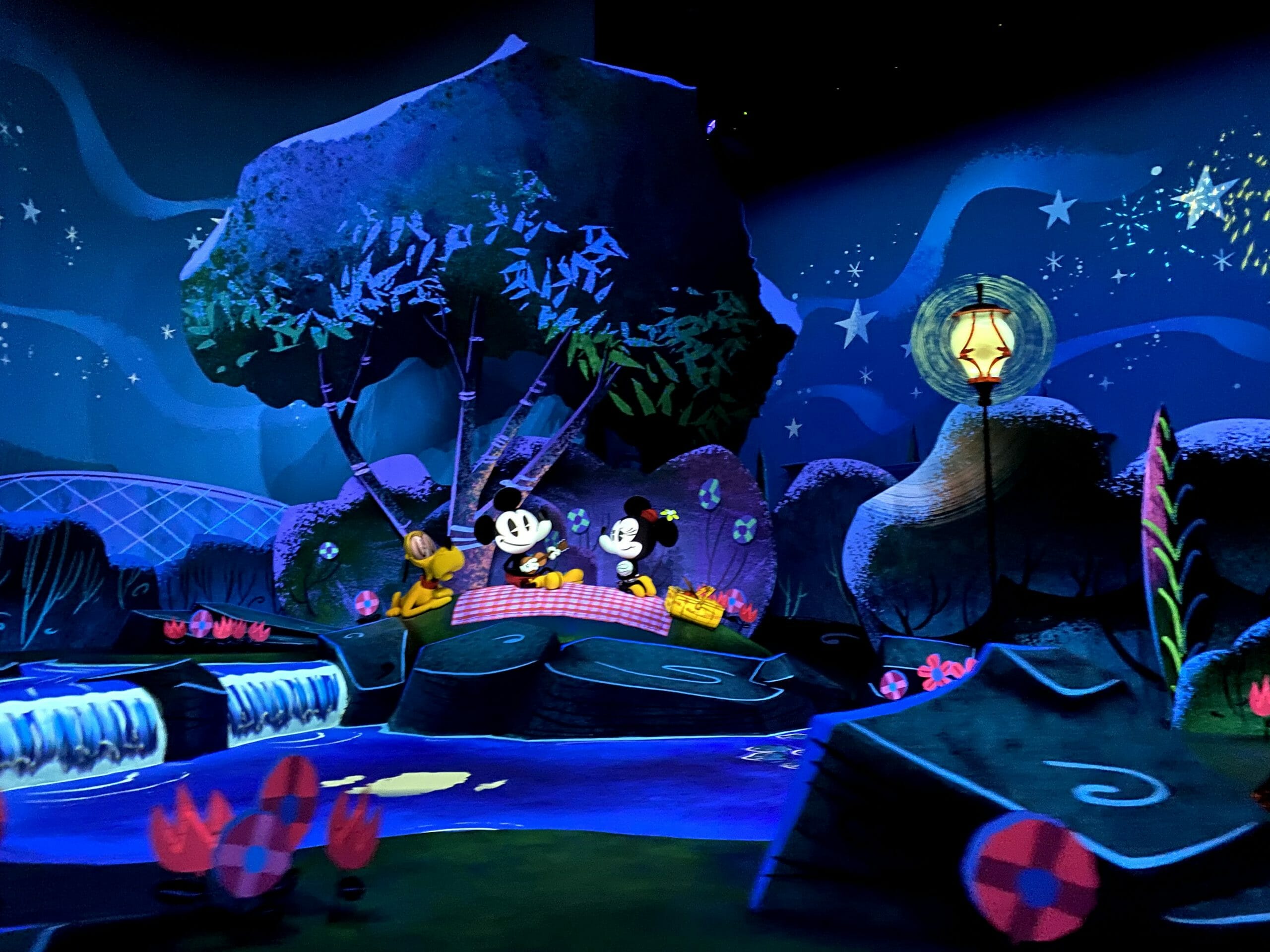 Imagem de divulgação da atração Mickey & Minnie's Runaway Railway, que mostra o Mickey, a Minnie e o Pluto fazendo um piquenique à noite sob árvores.