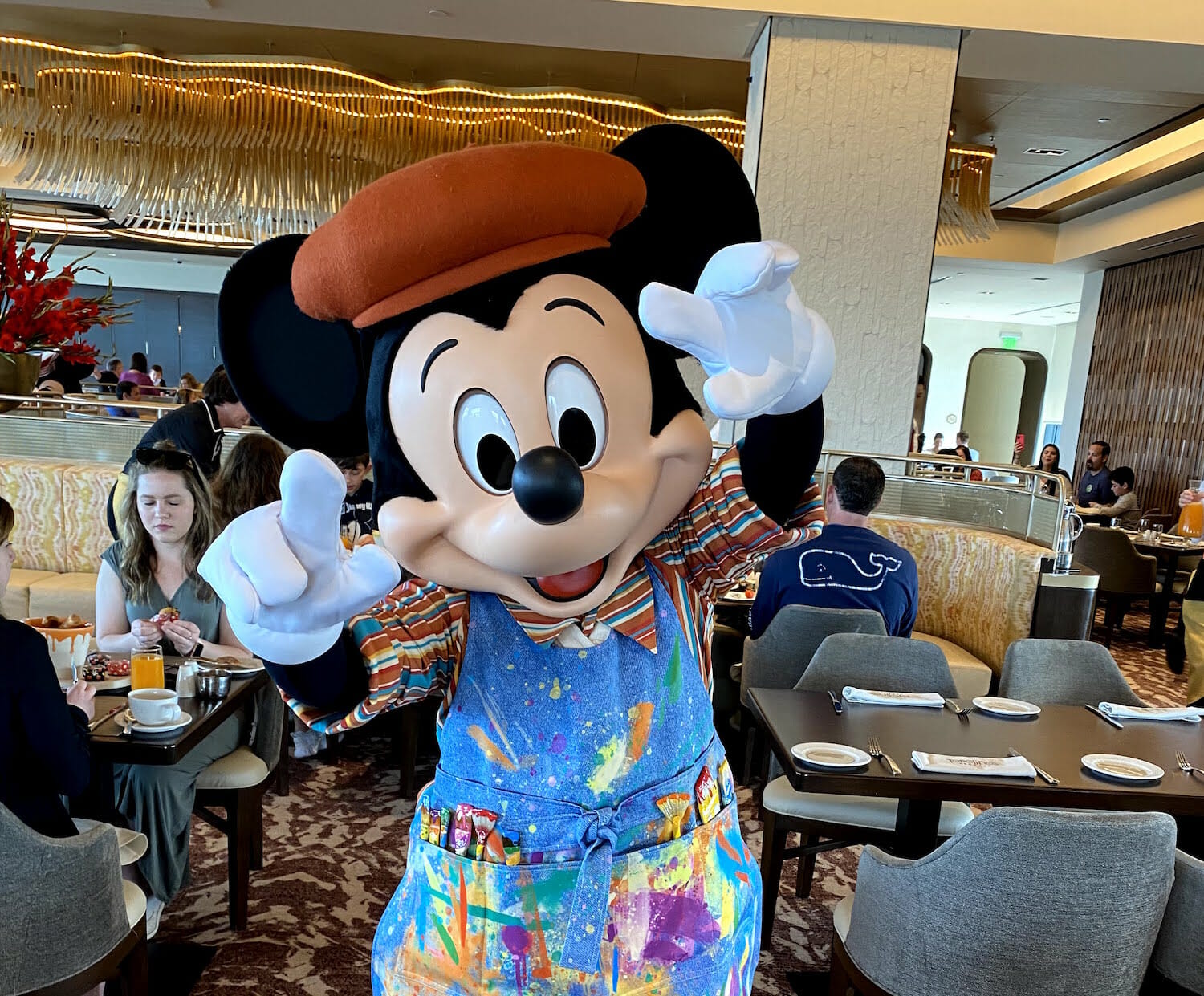 Foto do Mickey com o macacão de artista, manchado de tinta, e uma boina, a roupa com que ele aparece no café da manhã do Topolino's Terrace. Ele está acenando para a foto
