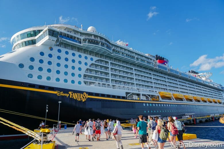 Imagem do navio de cruzeiro da Disney parado no porto, com visitantes em volta e o céu azul atrás. O navio é o Disney Fantasy, segundo as letras em amarelo escritas sobre ele.