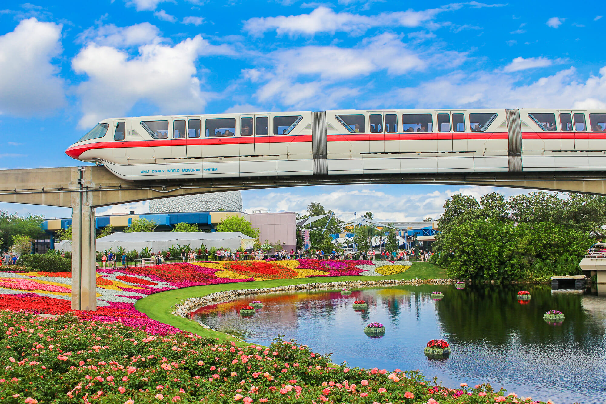 Foto do Monorail passando sobre o Epcot, na ponte em cima do lago, rodeada por flores e o céu azul ao fundo. Usada para ilustrar o post de wallpapers de celular