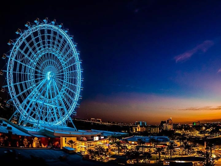 Foto da roda gigante The Wheel iluminada em azul, com o céu noturno ao fundo e a cidade de Orlando iluminada abaixo. 