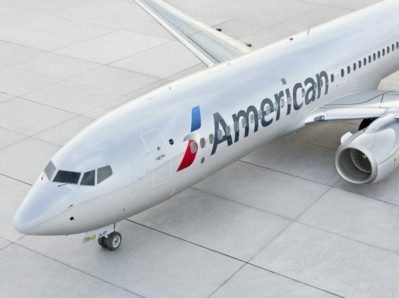 Imagem de um avião da American Airlines no aeroporto Imagem de um avião no aeroporto. Ele é branco, tem o logo azul e vermelho da American Airlines e a palavra "American" escrita em preto.
