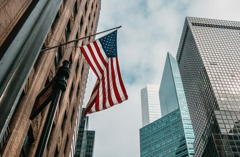 Foto de uma bandeira dos Estados Unidos balançando ao vento, com prédios modernos ao fundo.