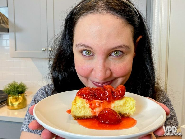 Foto da Renata segurando a fatia de cheesecake da Cheesecake Factory em frente à câmera e sorrindo. A cheesecake está coberta por morangos e calda de morango. 